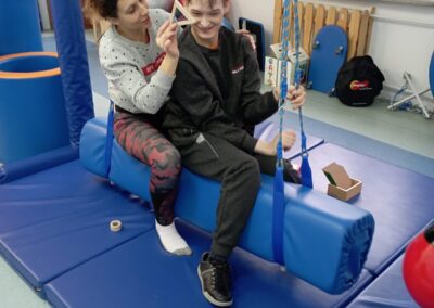 Nauczycielka siedzi za chłopcem na specjalnej huśtawce w pomieszczeniu do terapii sensorycznej. Oboje są uśmiechnięci, a nauczycielka pokazuje mu drewniany trójkąt.