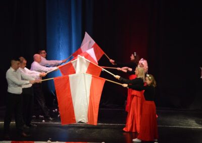 Grupa uczniów prezentująca patriotyczne flagi na scenie podczas występu. Flagi w biało-czerwonych barwach.