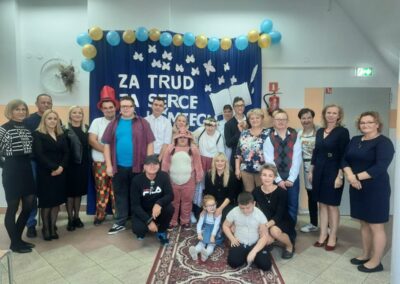 Grupa osób, w tym dzieci, ubrana w kolorowe stroje, pozująca do wspólnego zdjęcia. W tle znajduje się dekoracja z napisem "Za trud za serce".