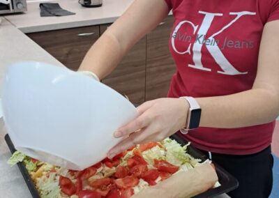 Uczennica w skupieniu przygotowująca potrawę, wsypując składniki do dużej formy na stole w klasie kuchennej.