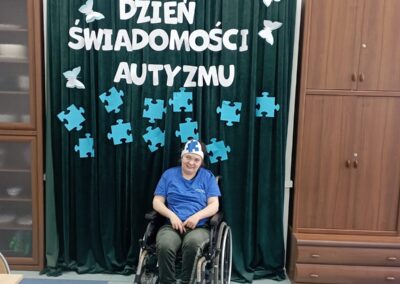 Kobieta na wózku inwalidzkim siedzi przed zieloną kotarą z napisem „Światowy Dzień Świadomości Autyzmu”. Dekoracja w tle składa się z niebieskich puzzli oraz białych motyli.