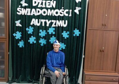 Mężczyzna na wózku inwalidzkim siedzi przed zieloną kotarą z napisem „Światowy Dzień Świadomości Autyzmu”. W tle widoczne są dekoracje w postaci niebieskich puzzli oraz białych motyli.