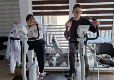 Dwie osoby ćwiczące na rowerach stacjonarnych w jasnym pomieszczeniu, z uśmiechem na twarzach. W tle duże okna zasłonięte roletami.