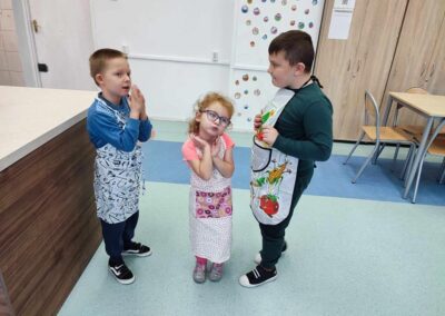 Trójka dzieci w fartuszkach stoi razem na środku pomieszczenia. Dziewczynka w różowej koszulce stoi pomiędzy dwoma chłopcami. Dzieci rozmawiają ze sobą, a w tle widoczna jest ściana z ozdobami i stół.