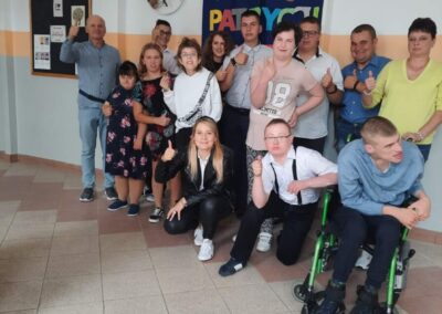 Grupa uczniów i nauczycieli świętująca urodziny w szkole, stojąca na tle ozdobionej ściany z napisem "Urodziny Kasi i Patrycji".