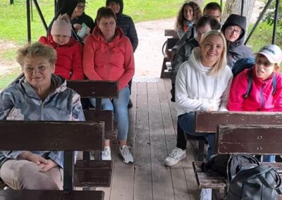 Grupa osób siedzi w drewnianym wozie konnym pod zielonym daszkiem, przygotowana na wycieczkę. Na pierwszym planie siedzi uśmiechnięta starsza kobieta.