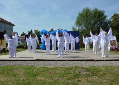 Grupa osób ubranych na biało podnosi ręce w geście zakończenia występu na scenie na świeżym powietrzu.