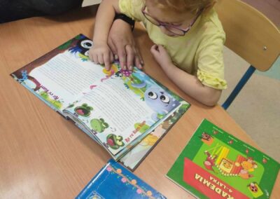 Dziewczynka w okularach i żółtej bluzce siedzi przy stole i czyta książkę, podczas gdy nauczycielka pomaga jej wskazując palcem na tekst. Na stole leżą jeszcze dwie inne książki. W tle widoczna jest drewniana szafka.