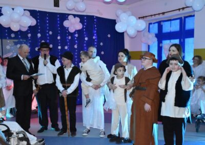Grupa osób przebranych za postacie z jasełek, w tym pasterze, anioły i inne postacie, stoją na scenie udekorowanej niebieskim tłem i białymi balonami w kształcie chmur. Dzieci w kostiumach są zaangażowane w przedstawienie.