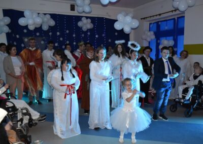 Kilka osób przebranych za anioły oraz królowie w kolorowych pelerynach stoją na scenie. Dziewczynka w białej sukni stoi na środku sceny.