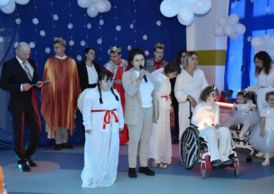 Aktorzy przebrani za anioły oraz postacie biblijne stoją na scenie, udekorowanej śnieżynkami i balonami. Kobieta z mikrofonem odgrywa rolę narratora, a obok niej znajdują się dzieci w strojach aniołów.