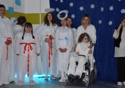 Grupa osób przebranych za anioły stoi na scenie. Dziewczynka na wózku inwalidzkim jest przebrana za anioła, obok inne osoby w białych szatach z aureolami na głowach.