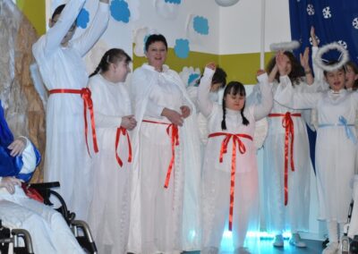 Kilka osób ubranych w białe stroje z czerwonymi wstążkami stoi na scenie, unosząc ręce. W tle niebieskie dekoracje z motywem śnieżynki. Całość nawiązuje do jasełek bożonarodzeniowych.