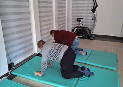 Dwóch chłopców klęczących na matach podczas ćwiczeń rehabilitacyjnych w jasnym pokoju z urządzeniami do ćwiczeń.