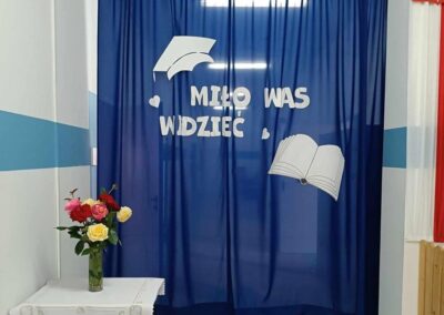 Niebieska kotara z dekoracją przedstawiającą napis Miło Was Widzieć, otwartą książkę oraz biret. Obok na stole stoi wazon z kolorowymi kwiatami.
