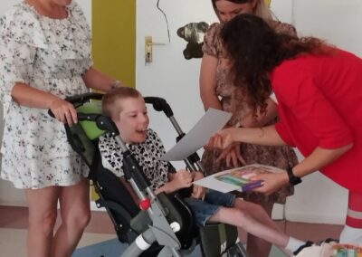 Dorośli współdziałają z dzieckiem na wózku inwalidzkim, pokazując mu rysunek w pomieszczeniu z dekoracją "MISIE".