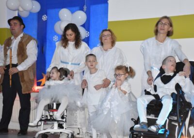 Kilka dzieci przebranych za anioły, w białych szatach, niektóre na wózkach inwalidzkich. Obok nich stoją dorośli opiekunowie. W tle dekoracje z balonami.