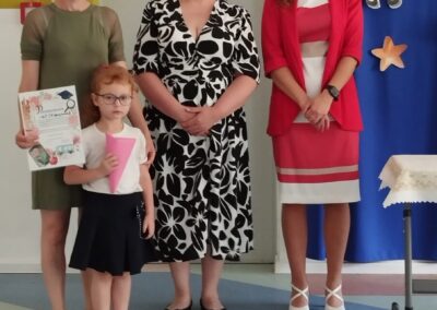 Dziecko z tekturowym stożkiem i dyplomem, pozujące z trzema dorosłymi na tle dekoracyjnej tablicy.