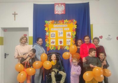 Grupa dzieci i opiekunowie z pomarańczowymi balonikami pozuje na tle plakatu Październik Miesiącem AAC. Dzieci i dorośli trzymają baloniki w kształcie piesków.