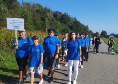 Grupa osób w niebieskich koszulkach idzie drogą podczas marszu. Jeden z mężczyzn trzyma tabliczkę z napisem 'Fundacja Szlachetne Anioły w Klimontowie'. W tle widoczny jest rowerzysta i zielone krzewy.