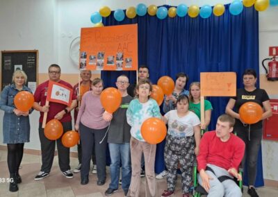 Grupa osób, w tym osoby z niepełnosprawnościami, pozuje z pomarańczowymi balonikami i plakatami na tle niebieskiej kurtyny. Jeden z plakatów ma napis Październik Miesiącem AAC.