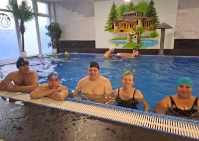 Grupa osób w basenie, stojących przy krawędzi basenu, przygotowujących się do pływania.