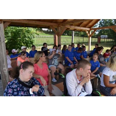 Duża grupa dzieci i dorosłych siedzi pod zadaszeniem na ławkach, większość dzieci ma na sobie niebieskie koszulki. W tle widoczna jest zieleń i ogrodzenie. Atmosfera jest relaksująca i spokojna.