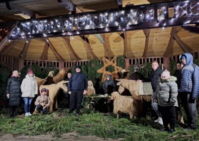 Grupa osób stoi przy szopce bożonarodzeniowej nocą, wokół postaci zwierząt wykonanych z naturalnych materiałów, podświetlonych lampkami.