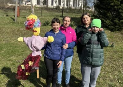 Grupa osób, w tym dzieci, stoi na trawie obok kolorowej kukły Marzanny. W tle widoczny kościół.
