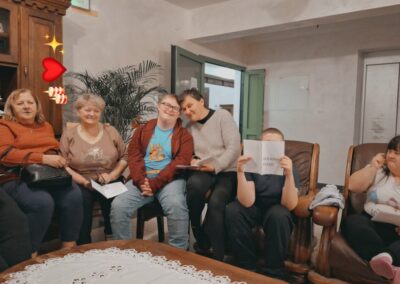 Grupa osób, w tym dzieci i dorośli, siedzi w sali i śpiewa kolędy, trzymając śpiewniki. Po lewej stronie widać dekoracje świąteczne.