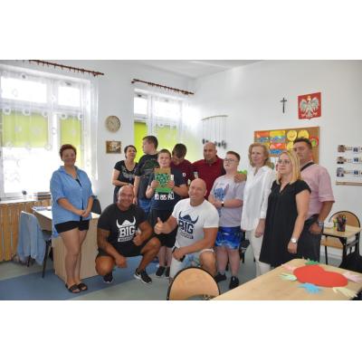 Grupa dzieci i dorosłych, w tym osoby z niepełnosprawnościami, pozuje w jasnej sali. W tle widoczne są dekoracje na ścianach, a w centrum dwóch mężczyzn w koszulkach z napisem „BIG” sugerujących gościnne wydarzenie. Niektóre osoby trzymają rękodzieła. Atmosfera jest przyjazna i wspierająca.