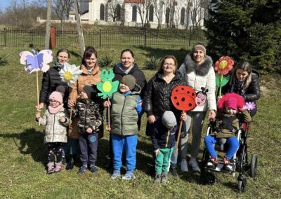 Grupa osób, w tym dzieci i dorośli, stoi na trawie z dekoracjami w kształcie motylka, biedronki, żaby i kwiatów. W tle widoczny kościół.