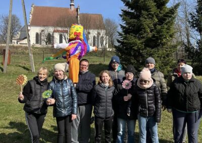 Grupa osób, w tym dzieci i dorośli, trzyma dekoracje i kukłę Marzanny na tle kościoła.