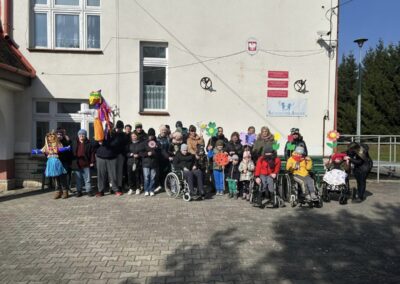 Grupa osób, w tym dzieci na wózkach inwalidzkich, stoi przed budynkiem szkolnym z dekoracjami i kukłą Marzanny.