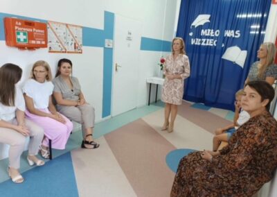 Grupa kobiet, w tym nauczycielki, rozmawia w szkolnym korytarzu na tle dekoracji Miło Was Widzieć. W tle siedzi chłopiec w białej koszulce.