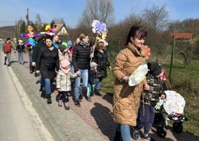 Grupa osób, w tym dzieci i dorośli, idzie chodnikiem przy drodze, trzymając kolorowe dekoracje. Część osób pcha wózki.