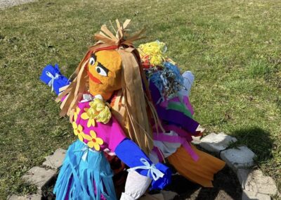 Kolorowa kukła Marzanny leży na trawniku, wokół niej kamienny okrąg. W tle widać dom.