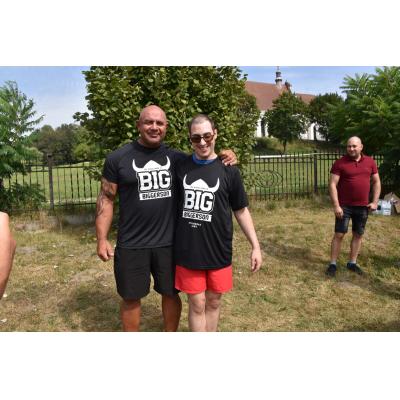 Dwaj mężczyźni w identycznych czarnych koszulkach z napisem „BIG BiggerSon” stoją na trawniku, jeden z nich obejmuje drugiego ramieniem. W tle widoczne jest ogrodzenie, drzewa oraz budynek przypominający kościół.