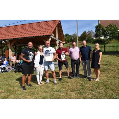 Grupa dorosłych pozuje w plenerze, trzymając nagrody i dyplomy. W tle widoczne są drzewa, trawnik oraz budynek. Atmosfera jest radosna i pełna uznania.