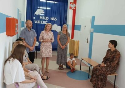 Grupa osób siedzi i rozmawia w szkolnym korytarzu przy dekoracji Miło Was Widzieć. Wśród uczestników są kobiety, mężczyzna i chłopiec.