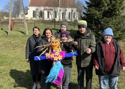 Grupa osób, w tym dzieci i dorośli, stoi na trawie obok kukły Marzanny. W tle drzewa i kościół.