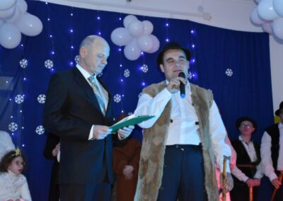 Mężczyzna w stroju pasterza trzyma mikrofon i przemawia. Obok niego elegancko ubrany mężczyzna trzyma kartkę. W tle dekoracje w postaci śnieżynek i światełek.
