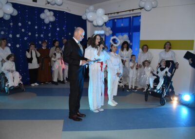 Mężczyzna w garniturze trzyma mikrofon, obok niego dziewczynka przebrana za anioła. W tle grupa postaci w strojach aniołów i pasterzy.