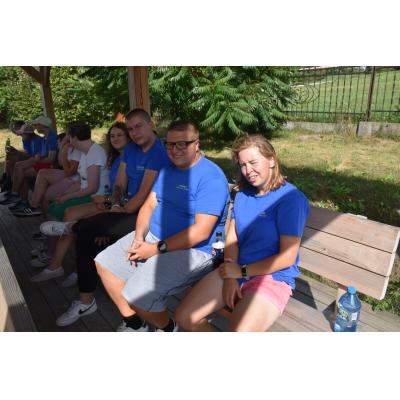 Grupa młodych dorosłych siedzi na ławkach pod zadaszeniem na świeżym powietrzu. Wszyscy noszą niebieskie koszulki. W tle widoczne jest ogrodzenie i zieleń. Atmosfera jest spokojna i relaksująca.