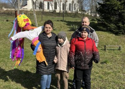Rodzina z dziećmi stoi na trawie obok kukły Marzanny. W tle widoczny kościół i drzewa.