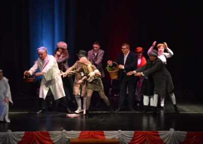 Uczestnicy występu na scenie trzymający kosze, ubrani w kostiumy, podczas przedstawienia teatralnego.