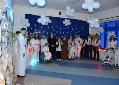Grupa osób w różnych strojach, anioły, pasterze i inne postacie stoją na scenie. Tło ozdobione śnieżynkami i światełkami.