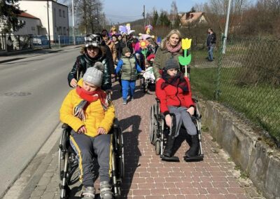 Grupa osób, w tym dzieci na wózkach inwalidzkich, idzie chodnikiem. W tle widoczne osoby z kolorowymi dekoracjami.