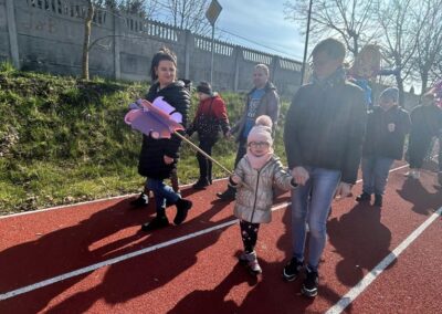 Grupa osób, w tym dorosłe kobiety i dziecko, idzie po bieżni na świeżym powietrzu, trzymając w rękach kolorowe rekwizyty. W tle drzewa i mur.
