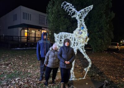 Trójka dzieci stojąca przed dużą świetlną instalacją w kształcie renifera podczas nocnego spaceru.